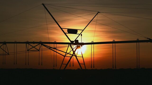 Silhouette of center-pivot irrigation system against vibrant orange sunset sky in vast agricultural field, showcasing modern farming technology and water management during golden hour