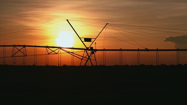 Silhouette of a center pivot irrigation system standing in a vast agricultural field during a vibrant orange sunset, highlighting modern farming technology and water management at dusk