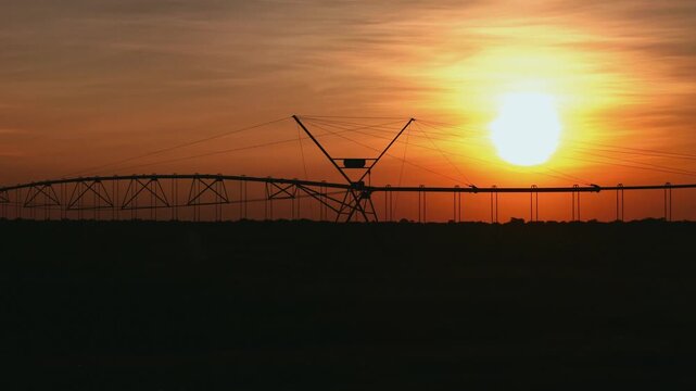 A center pivot irrigation system stands silhouetted against a vibrant orange sunset over a vast agricultural field, highlighting modern farming technology and rural landscape beauty at dusk