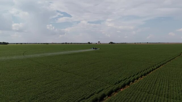 Aerial view of a professional crop duster airplane flying low over an expansive green agricultural field, releasing a trail of spray to protect crops under a bright, cloudy summer sky