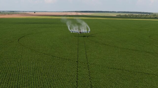 Aerial view of an agricultural crop duster airplane flying low over a lush green field, spraying liquid pesticides or fertilizers to protect crops in a vast rural farming landscape