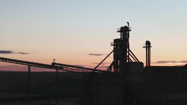 Silhouette of a massive industrial grain elevator and conveyor belt infrastructure against a soft sunset sky in a rural landscape, highlighting agricultural logistics and heavy machinery at dusk