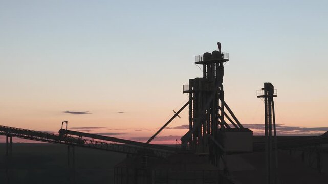 Silhouette of a large industrial grain elevator and conveyor belt system against a colorful sunset sky at dusk, highlighting agricultural infrastructure and rural landscape during golden hour