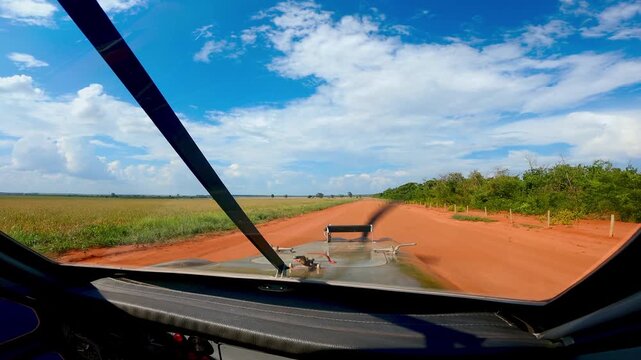 First-person perspective view from the cockpit of a heavy vehicle or armored transport driving along a red dirt road through lush green agricultural fields under a bright blue sky with clouds