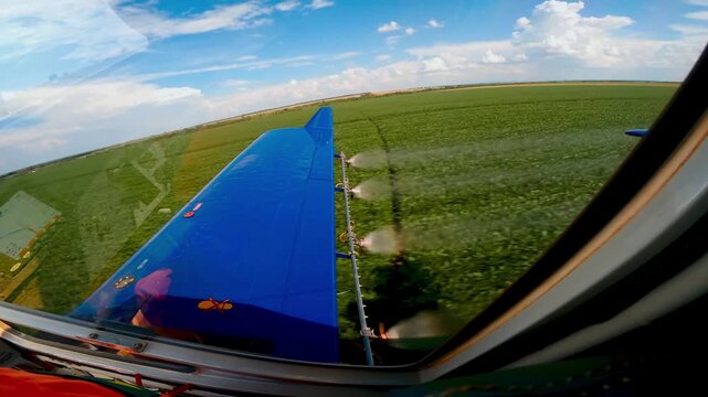 Agricultural aircraft wing spraying pesticide over vast green farm fields under a blue sky with white clouds, capturing the perspective of a crop duster pilot during a low-altitude flight