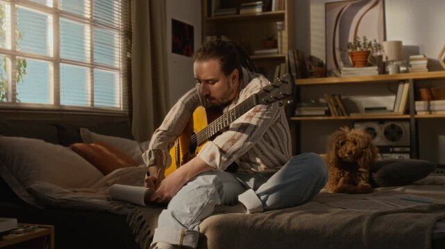 Caucasian young male artist playing acoustic guitar and writing song lyrics in notebook while sitting on bed next to small pet dog in home studio. Music education, freelance and creative hobby ads