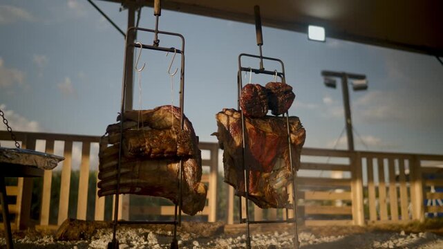 Traditional Argentine asado featuring large cuts of beef roasting on vertical metal racks over an open fire outdoors under a blue sky at a rustic ranch or cultural food festival event