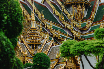 The ornate golden roof of a temple in The Grand Palace, Bangkok, Thailand, is framed by lush green foliage, showcasing intricate architectural details. © Zenstratus