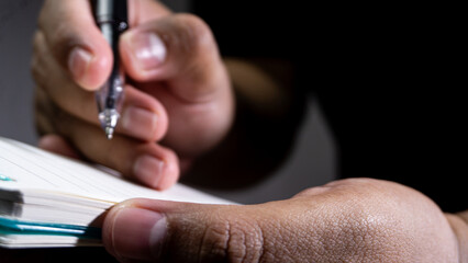 Close up of a male hand holding a black pen and a notebook, ready to write on a lined page, with visible skin texture, shadows, and dim lighting against a dark grey background. © M. Faisal Riza