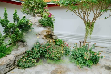 Fotobehang Badkamer A serene garden with a mist effect cascading over rocks and blooming roses, located at Wat Pho, Bangkok, Thailand. Atmosphere is enhanced by the lush greenery and delicate flowers.  © Zenstratus