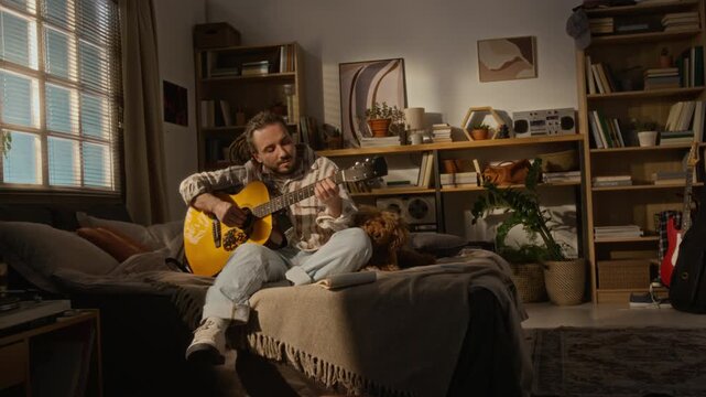 Caucasian young man sitting on bed, playing acoustic guitar next to small dog in home studio. Footage ideal for promoting creative hobbies, remote lifestyle, freelance work or pet companionship