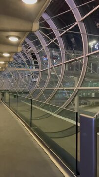 Vertical POV walking through a modern illuminated glass tube tunnel and moving walkway in an empty airport terminal at night.