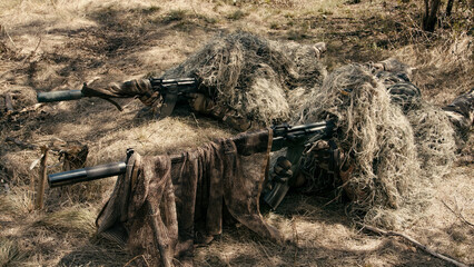 Naklejka premium Sniper team camouflaged with ghillie suits lying on the ground in a forested area, rifles positioned for a tactical mission in a natural environment
