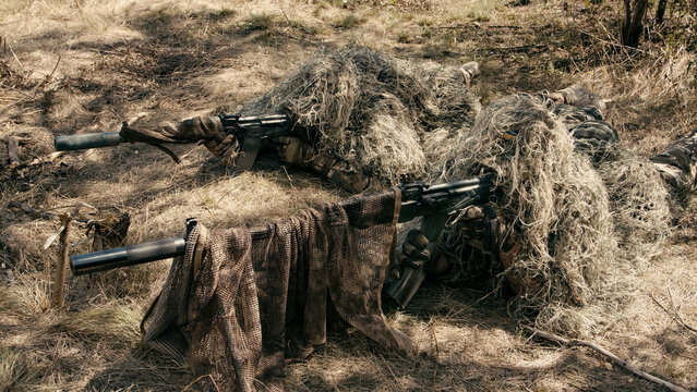 Sniper team camouflaged with ghillie suits lying on the ground in a forested area, rifles positioned for a tactical mission in a natural environment