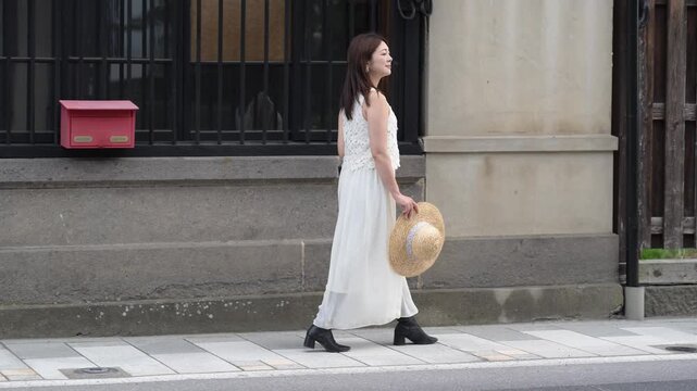 白いワンピースを着て麦わら帽子を持ち古い町並みを散策する女性（夏の一人旅）