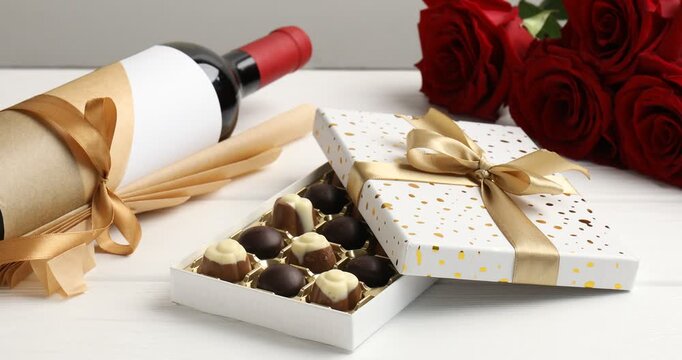 Putting red roses onto white wooden table with bottle of wine and box with candies, closeup
