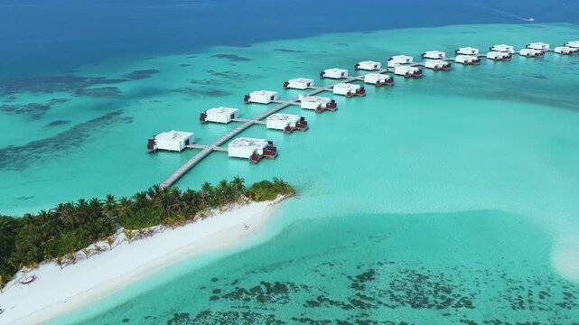 Aerial view of luxury overwater bungalows extending into the turquoise lagoon from a tropical island with palm trees in Kudahuvadhoo, Dhaalu Atoll, Maldives.