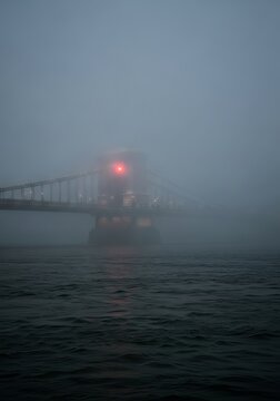 Suspension bridge shrouded in thick fog over dark river water. Glowing red warning light on stone pylon during gloomy evening. Mysterious atmospheric city scape with urban architecture.