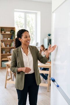 Smiling Female Teacher Gesturing Towards Whiteboard While Teaching Students in a Bright Modern Classroom