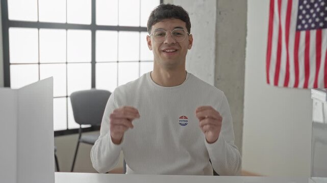 Man smiling and gesturing with hands, wearing a voted sticker while seated at a polling building desk; civic pride.