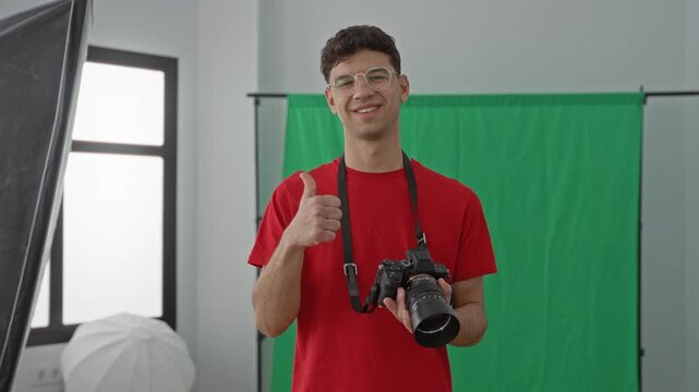 Young man photographer holding camera and giving thumbs up in studio with green screen and softbox lights, red shirt and glasses; creative confidence.