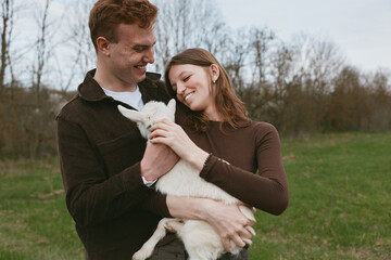 Obraz premium Joyful couple outdoors, holding a fluffy goat kid, warm affection, bright sky, casual attire, grassy field, natural colors, candid moment, rustic charm, celebration of pet companionship