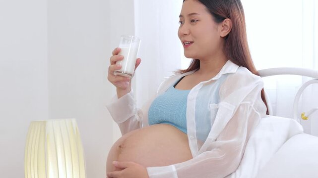 Close-up of a pregnant mother drinking milk for calcium nutrition. Smiling pregnant woman holding milk glass on bed, taking care of her unborn baby health.