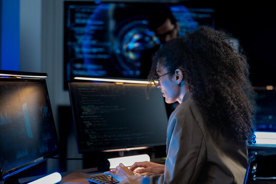 South Asian male and Black female software developers pair-program at dual monitors displaying network security algorithms at night.