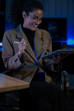 Black female developer works alone at a laptop in a dark tech office, lit only by the glow of the monitors around her.
