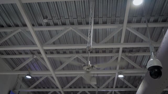 Handheld shot captures a large industrial fan in motion inside a warehouse, its spinning blades filling the frame against a gray ceiling structure background in a clean neutral industrial setting