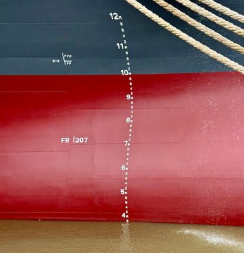 Close-up view of a container ship hull showing draft marks and mooring lines alongside the vessel. Maritime transportation details captured from ship&rsquo;s side with industrial nautical elements visible
