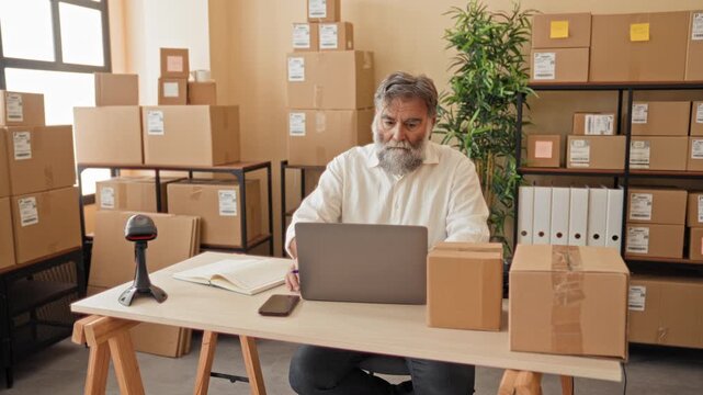 Man with gray beard writing notes at a laptop among parcel boxes in a packing area inside a building; diligence entrepreneurship.