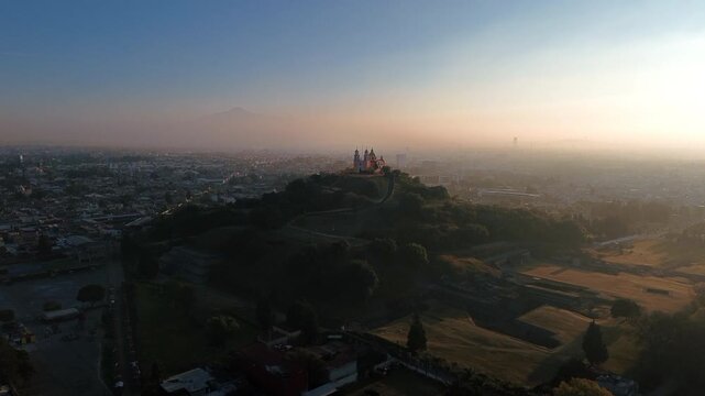 Aerial drone moves cinematically close to the Church of Our Lady of Remedies atop the Cholula pyramid at dawn, with the town and Puebla city spreading across the background