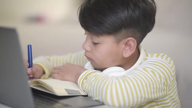 Focused Asian schoolboy writing notes in workbook while studying with laptop. Concentrated preteen pupil engaged in online education course and doing digital homework