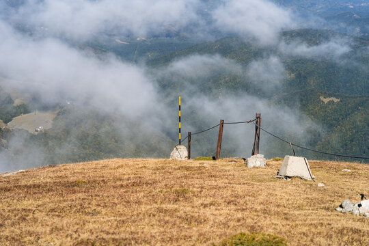 View of Botev Peak with a metal fence and marker post on the grassy summit, surrounded by clouds and mountain slopes in Balkan Mountains, Lovech, Bulgaria.