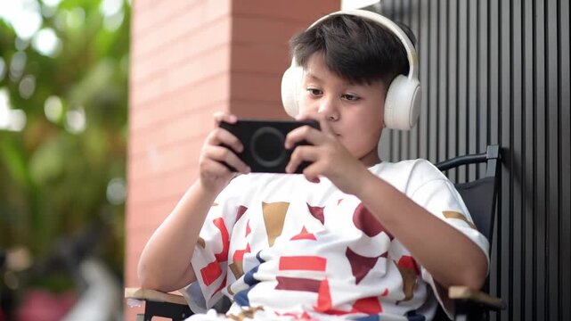 Cheerful Asian schoolboy wearing wireless headphones interacting with smartphone outdoors and cheering. Excited preteen pupil successfully winning gamified edtech course
