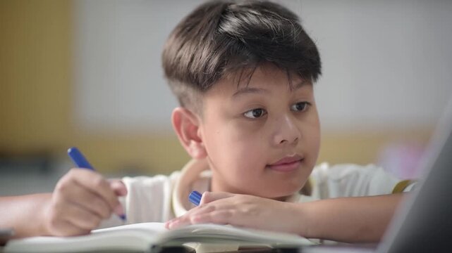 Focused Asian schoolboy writing notes in workbook while studying digital online education course. Concentrated preteen pupil engaging with modern distance learning class
