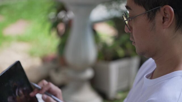 Handheld swaying shot of focused Asian man face working with stylus and blurred tablet in green garden. Organic camera movement footage for modern remote workflow concepts.
