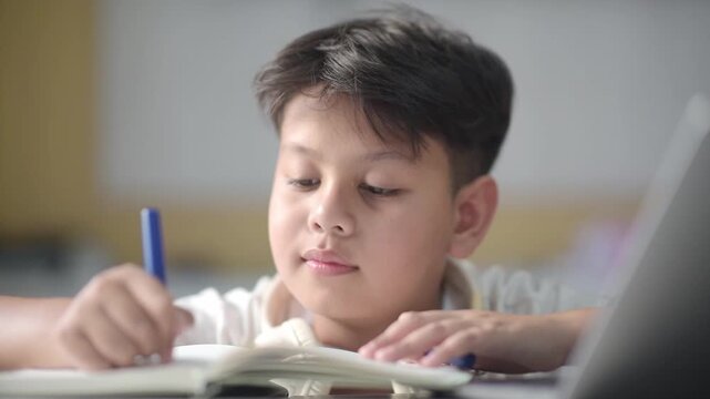 Smart Asian schoolboy writing notes and thinking deeply before smiling brightly with new realization. Clever preteen pupil successfully solving difficult academic assignment