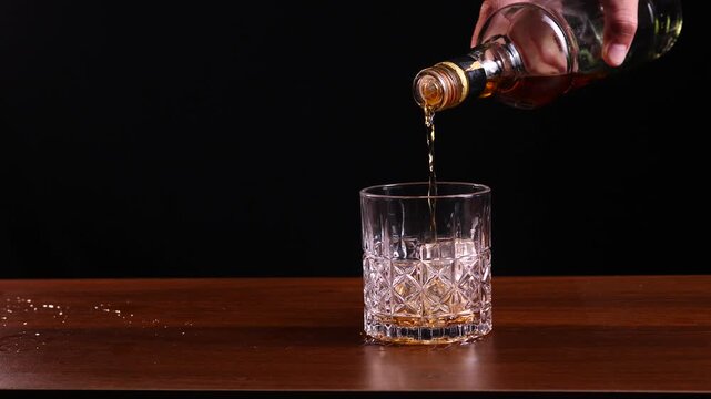 Pouring whiskey from a bottle into a patterned crystal glass on a wooden table with a black background