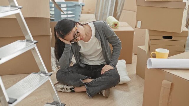 Woman sitting crosslegged among stacked cardboard boxes and a ladder, hand to head while unpacking in apartment; weary relocation stress.