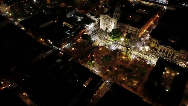 Aerial drone night view of historic city square filled with large crowd surrounding illuminated cathedral and vibrant urban center during public gathering