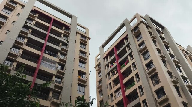 A low angle pan shot of contemporary highrise apartment complexes in India, featuring balconies and red architectural accents under a cloudy sky.