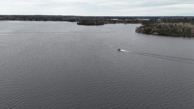 Lake of the Woods near Kenora, Ontario. Aerial drone ascends above open water as two boats cross the frame, with distant islands visible under a cloudy spring sky.