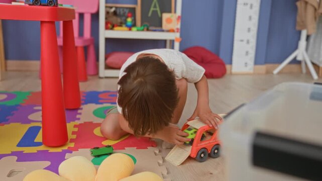 Boy kneeling with hands on toy truck and small green car, reaching across colorful foam puzzle mat in building playroom; childhood joy.