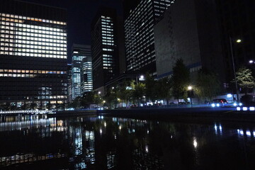 夜の都市景観。明るく照らされた高層ビルが河面に映り込み、緑の木々と柔らかな街灯が周囲に広がる静かな夜景。 © BRUCE