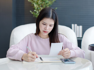 Naklejka premium Asian happy beautiful woman working writing record information on paper sitting on modern living room ,lifestyle enjoy wellbeing healthy young person relaxed mood peaceful security wellness at home.