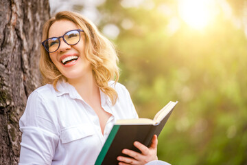 Fototapeta premium Laughing woman student in glasses reading book in park. Lifelong learning and joy of education concept.