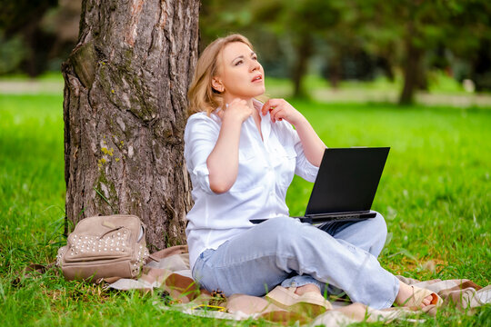 Tired woman freelancer adjusting shirt working on laptop near tree. Remote work difficulties and heat wave discomfort concept.