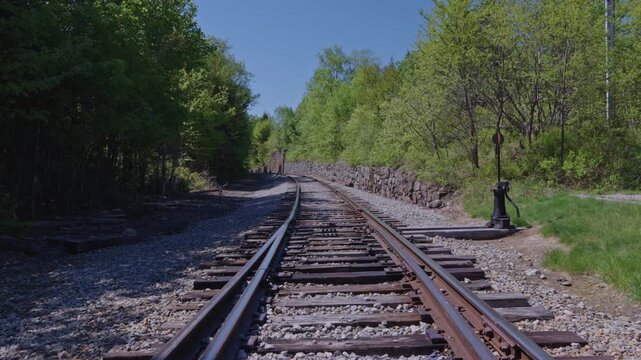 Empty train tracks curving past stone wall and switch stand into green and sunny landscape